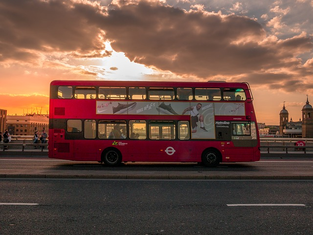 A red double decker bus is driving down a London street at sunset.