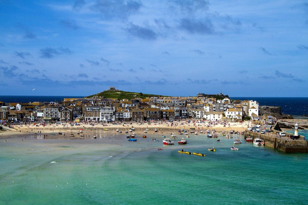 An aerial view of a beach in St Ives with boats in the water and a lighthouse in the distance.
