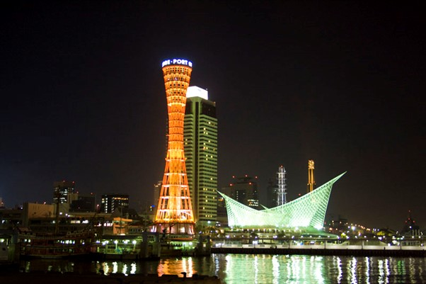 the kobe skyline at night with a tower in the foreground and a boat in the background.