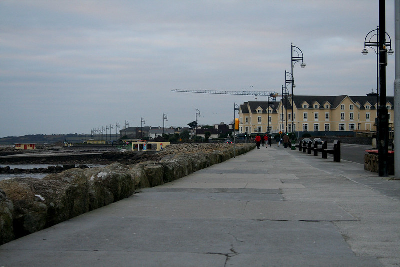 A concrete walkway along the Galway seafront with a large building in the background.