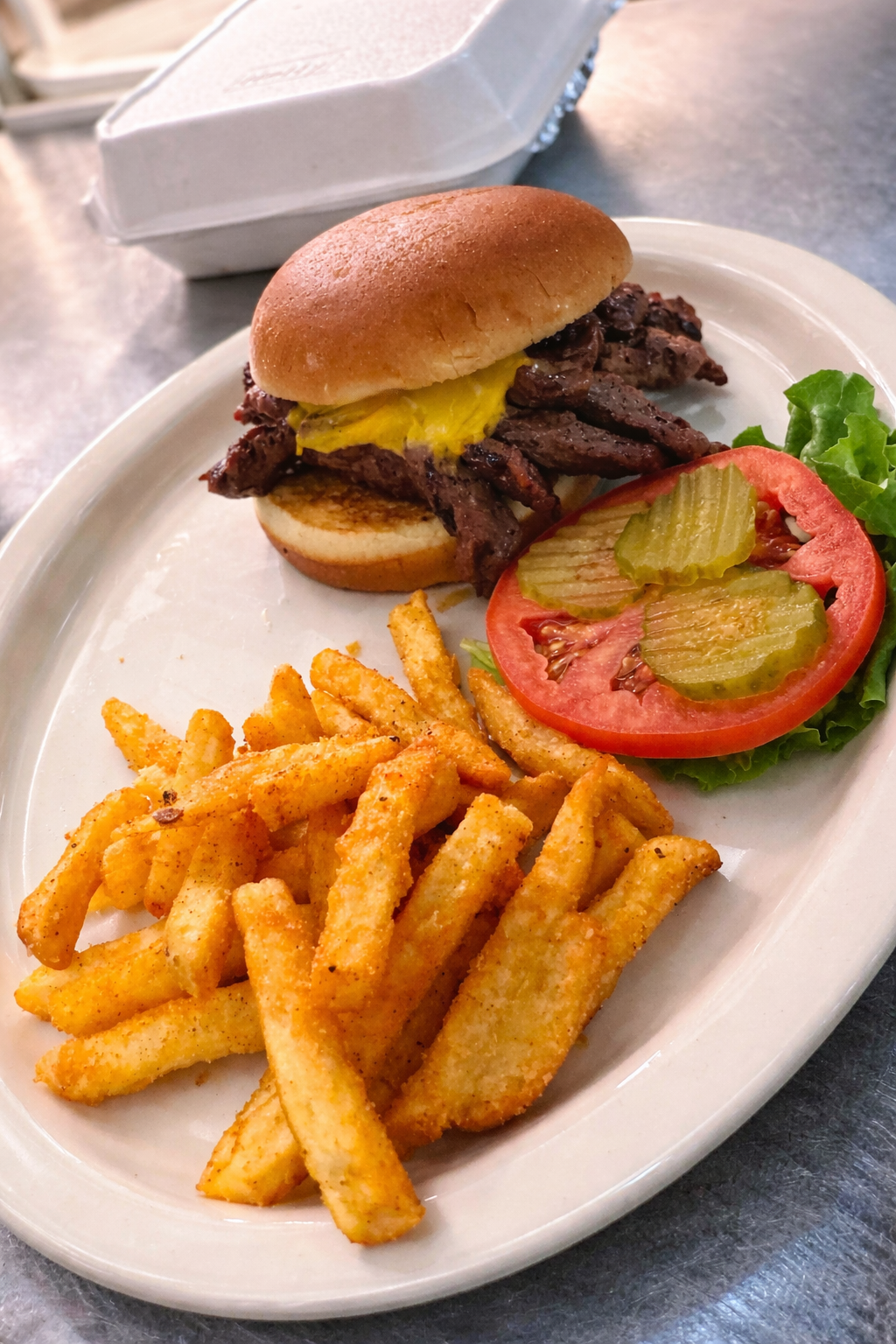 A steak sandwich on a bun with melted cheese, served with fries, a tomato slice, pickles, and lettuce on a white plate.