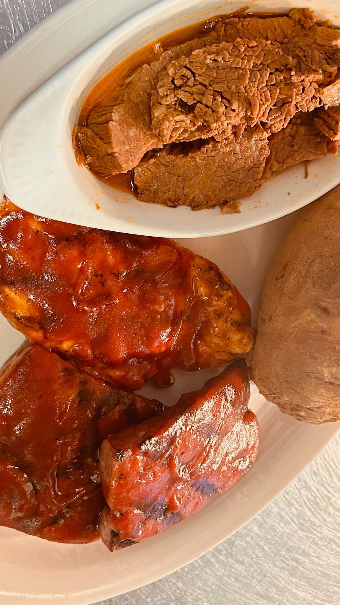 A plate featuring a portion of ribs covered in red sauce, a side of brown mashed food, and a whole baked potato.