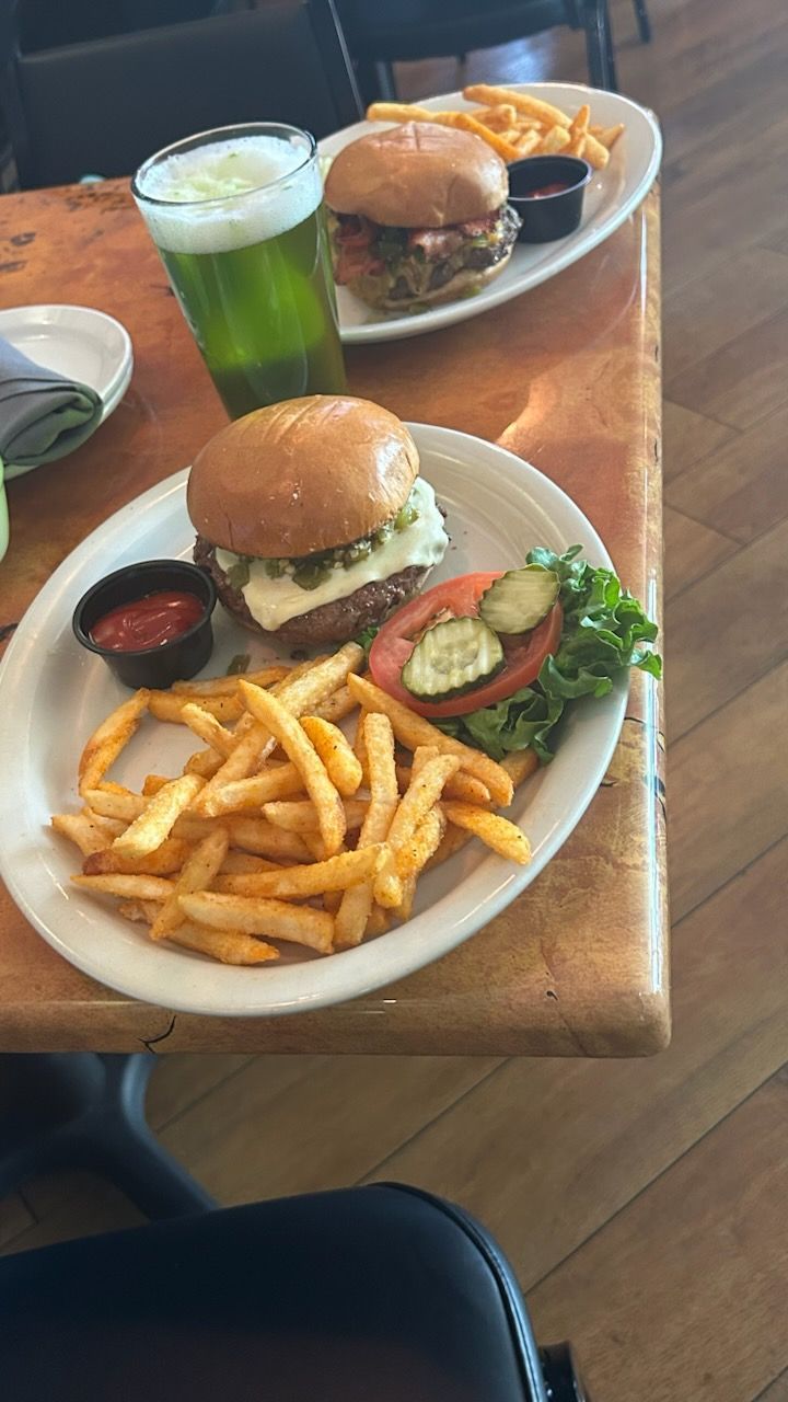 Two burger meals with French fries and dipping sauce on a wooden table, accompanied by a glass of green liquid.