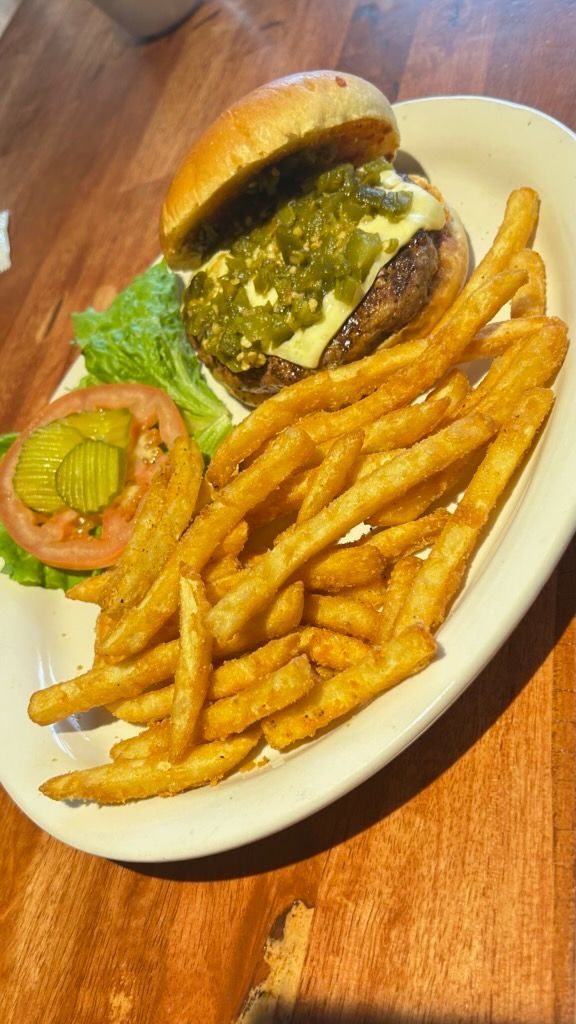 A burger topped with green chiles and cheese, served with a side of fries, lettuce, tomato, and pickles on a white plate.