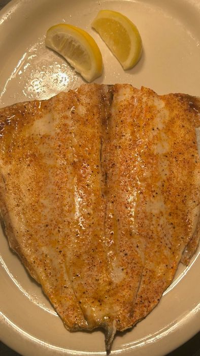 A seasoned fish fillet, cooked and drizzled with oil, served on a white plate with two lemon wedges.