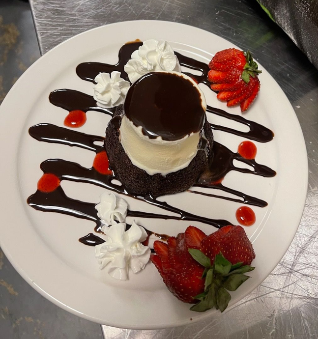 A chocolate lava cake topped with vanilla ice cream, drizzled with chocolate sauce, whipped cream, and fresh strawberries.