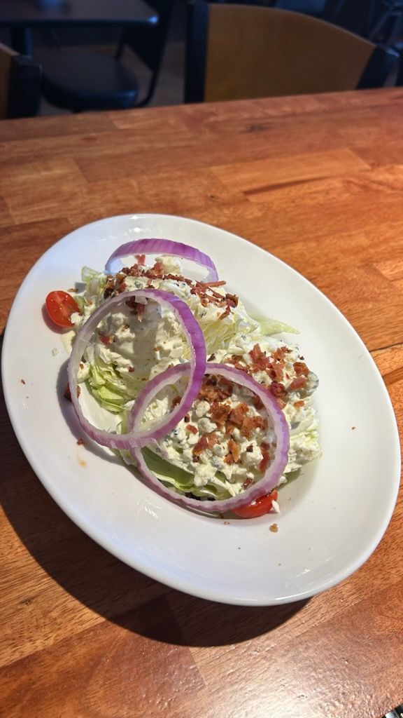 A wedge salad topped with blue cheese dressing, bacon bits, red onion rings, and cherry tomatoes on a white oval plate.
