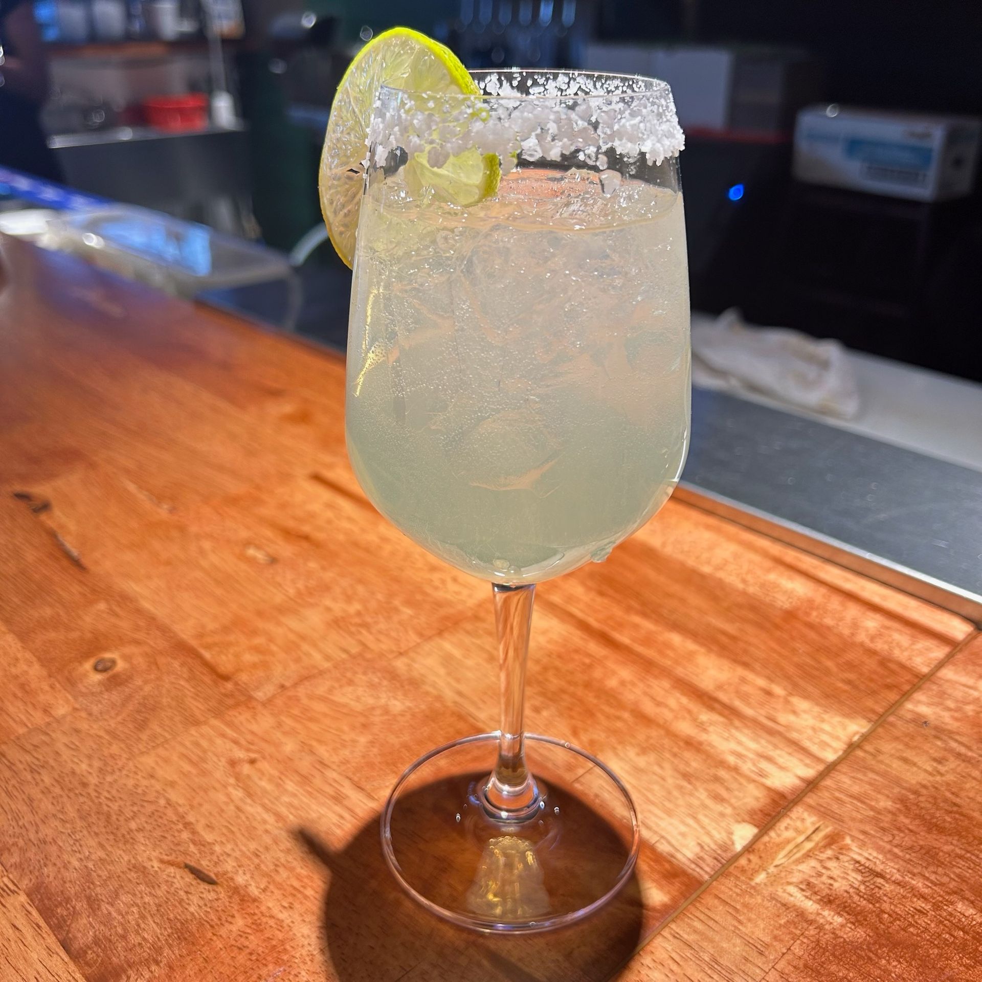 A chilled, salt-rimmed margarita in a stemmed glass with a lime wedge garnish, sitting on a wooden bar counter.