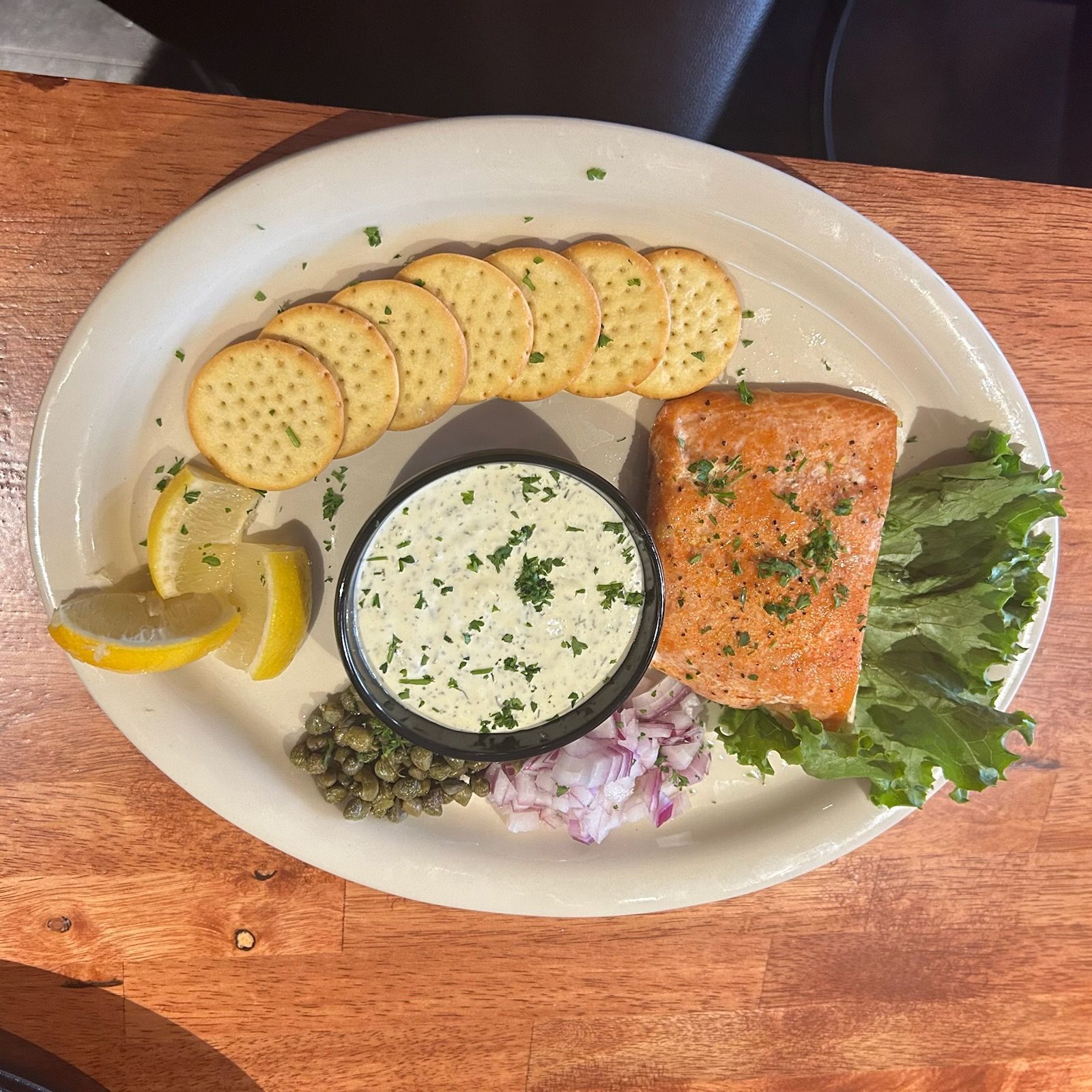 A plate featuring a salmon fillet, a bowl of herb dip, round crackers, red onion, capers, lettuce, and lemon wedges.