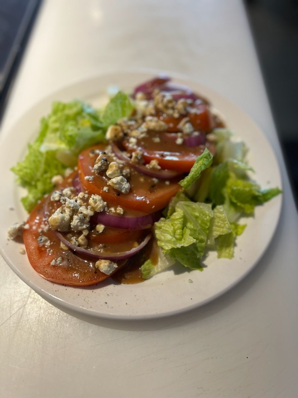 A salad with sliced tomatoes, red onions, blue cheese crumbles, and dressing, served on a white plate.
