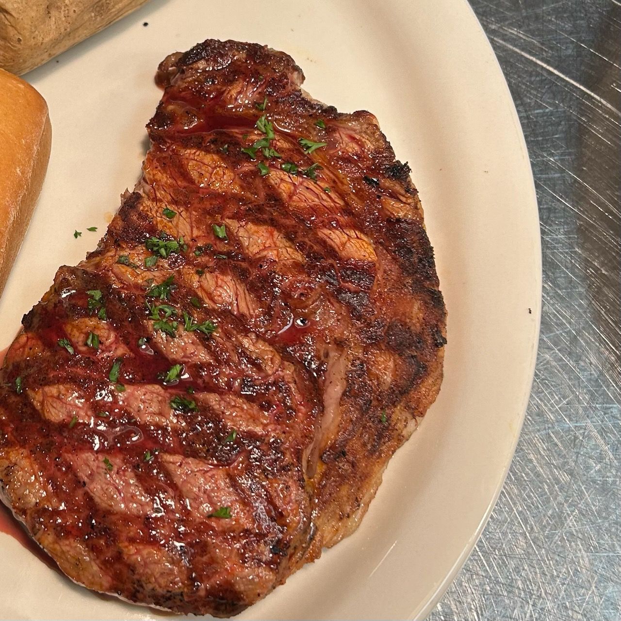 A grilled ribeye steak with char marks and a herb garnish, served on a white plate next to a piece of bread.