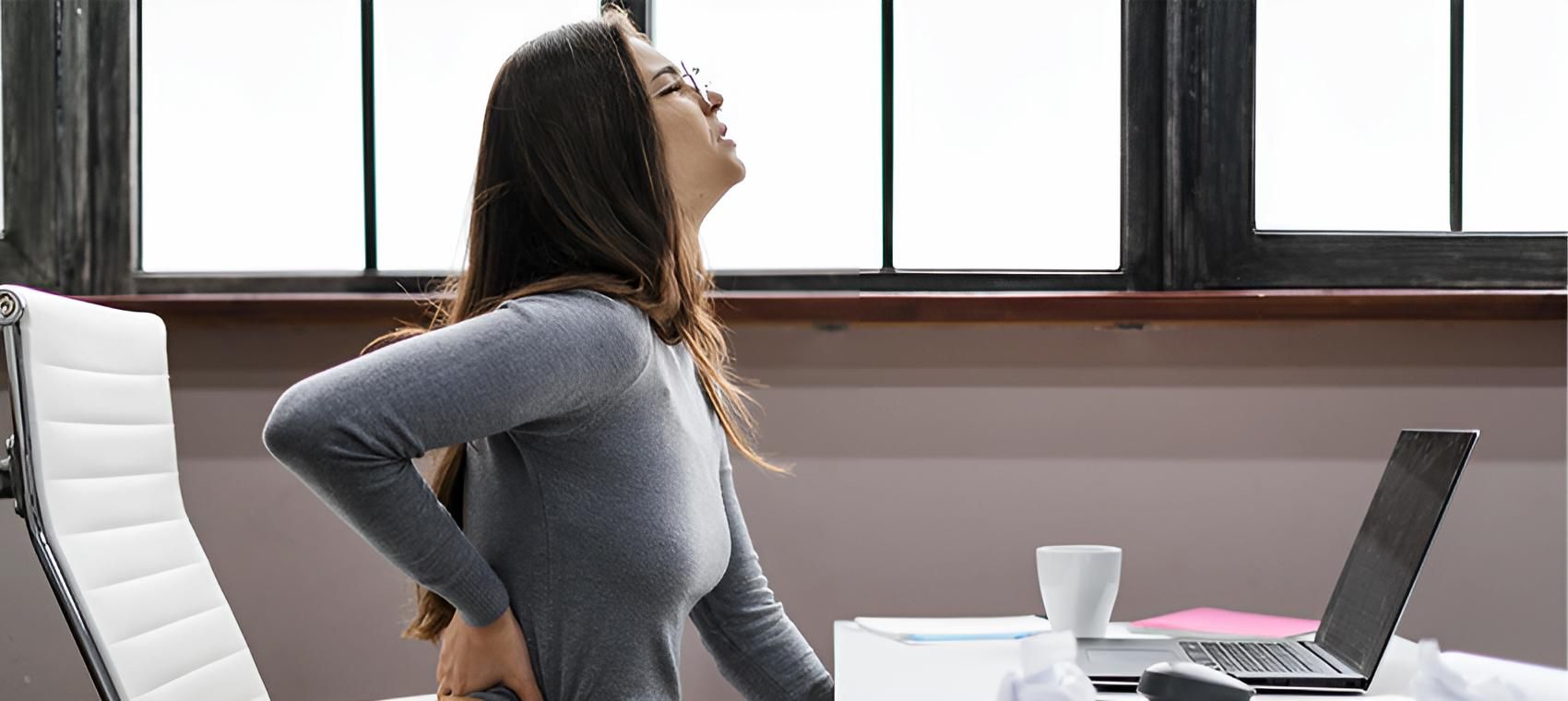A Woman is Sitting at a Desk With a Laptop and Holding Her Back — Back Care Gold Coast in Nerang, QLD