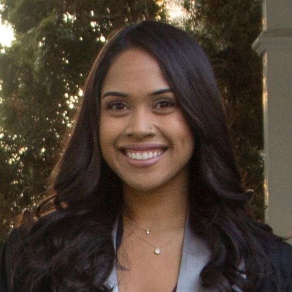 A smiling person with long brown hair, wearing a white top and a blue graduation stole, poses outdoors by a tree.