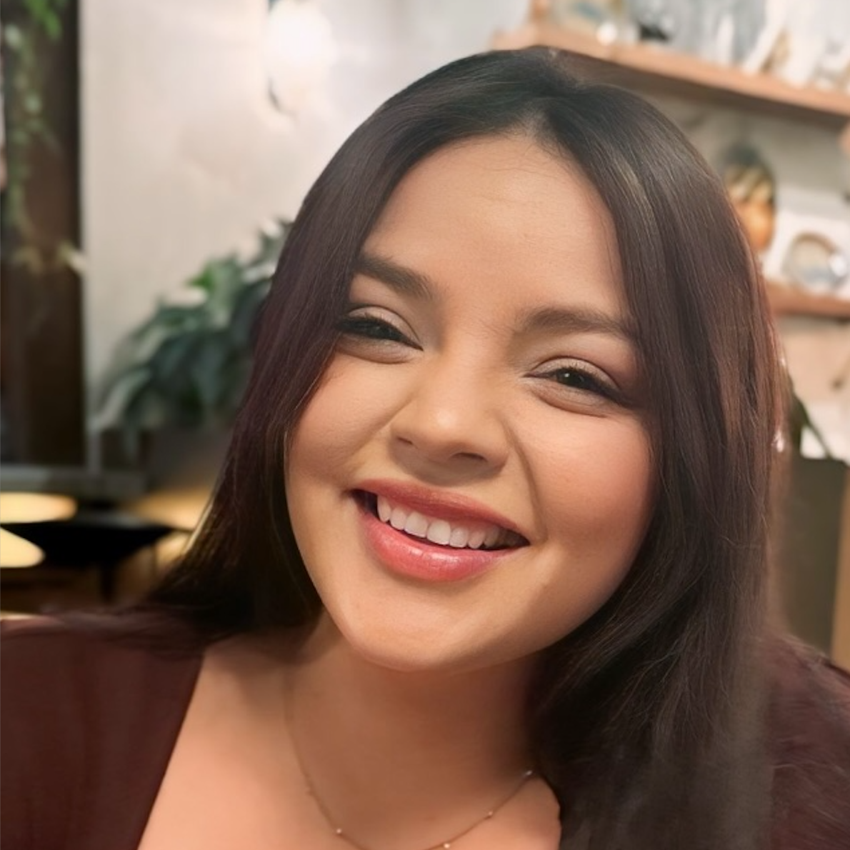 A person with long brown hair smiles broadly while wearing a dark top, centered in a softly lit, indoor setting.