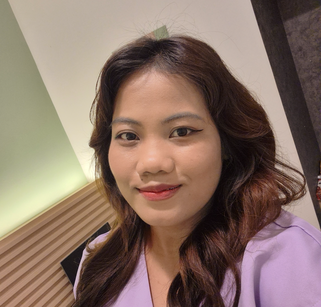 A person with dark hair smiling at the camera, wearing a lavender top in an indoor room with a wood-paneled wall.