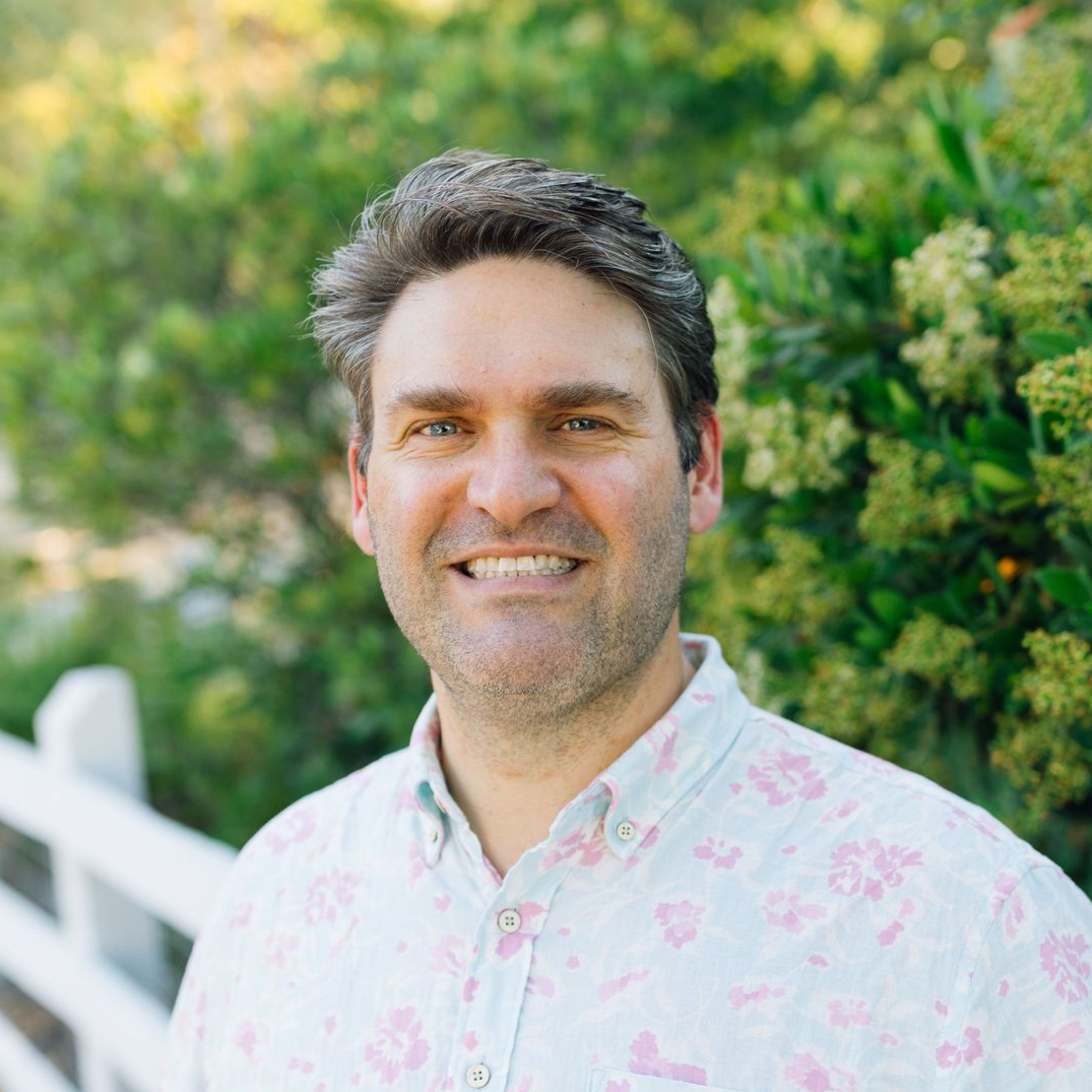 A smiling person with dark hair wears a light-colored, patterned button-down shirt outdoors near a white fence and bushes.