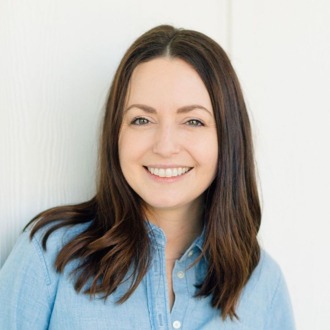 A smiling person with long dark hair wearing a light blue denim shirt against a plain white background.