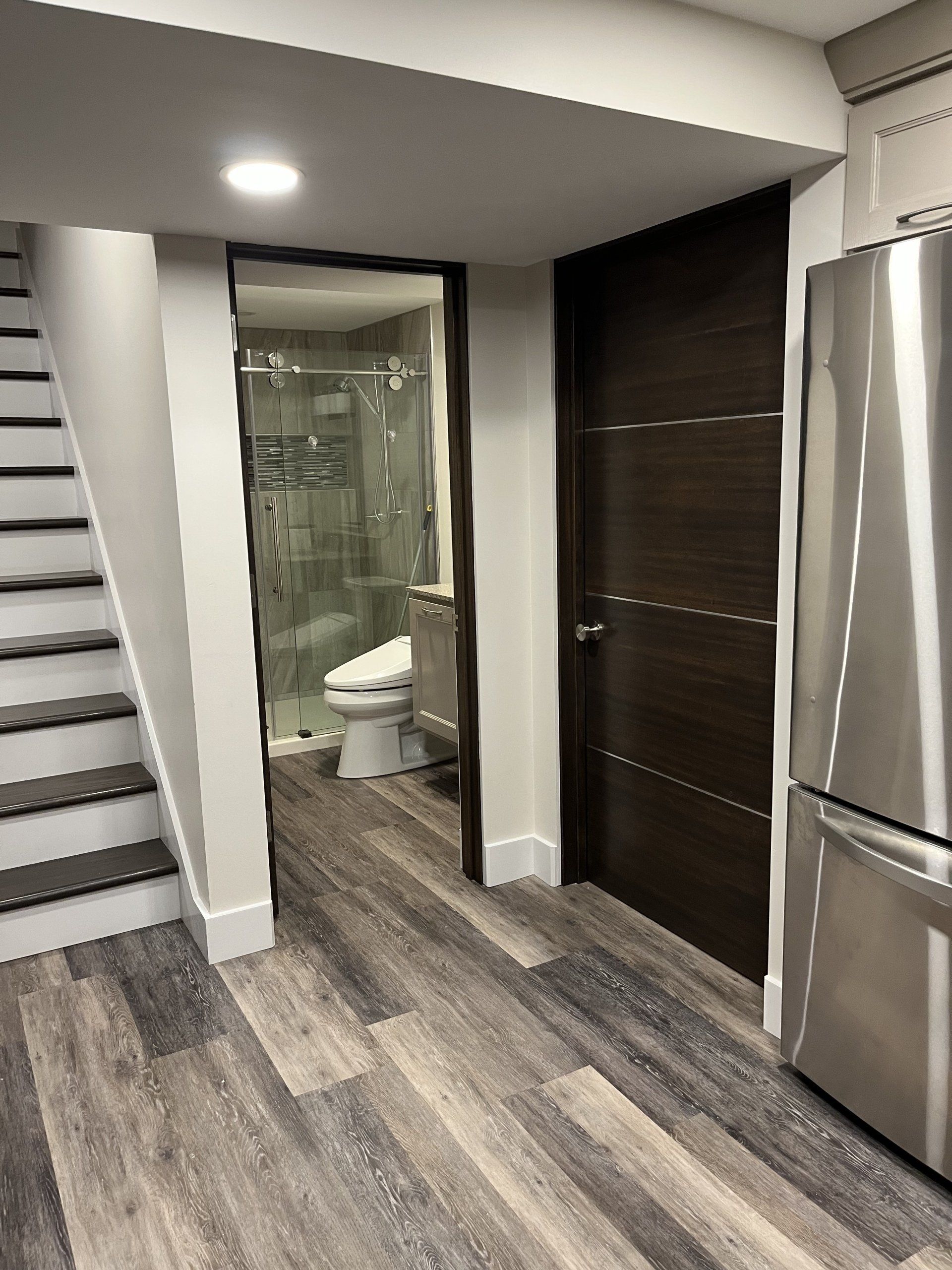 Modern hallway with dark wood floor, stainless fridge on right, stairs on left, and a bathroom visible ahead