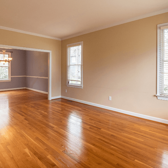 Empty beige living room with hardwood floors, white trim, and two windows overlooking an adjoining room