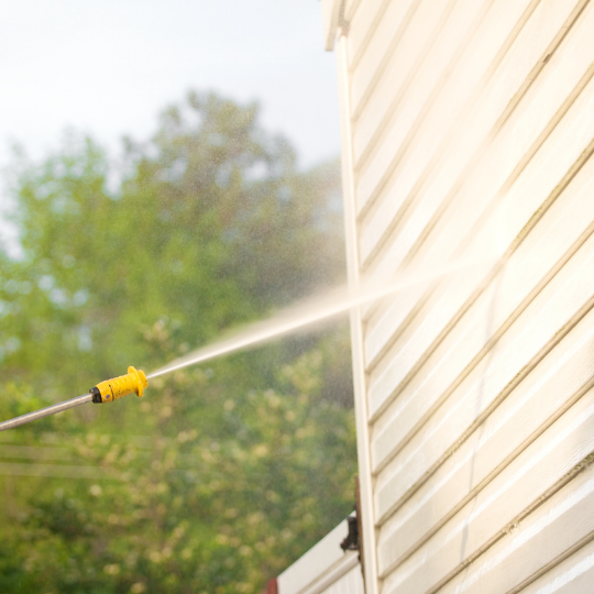 A hose spraying water at a beige house siding with trees in the background