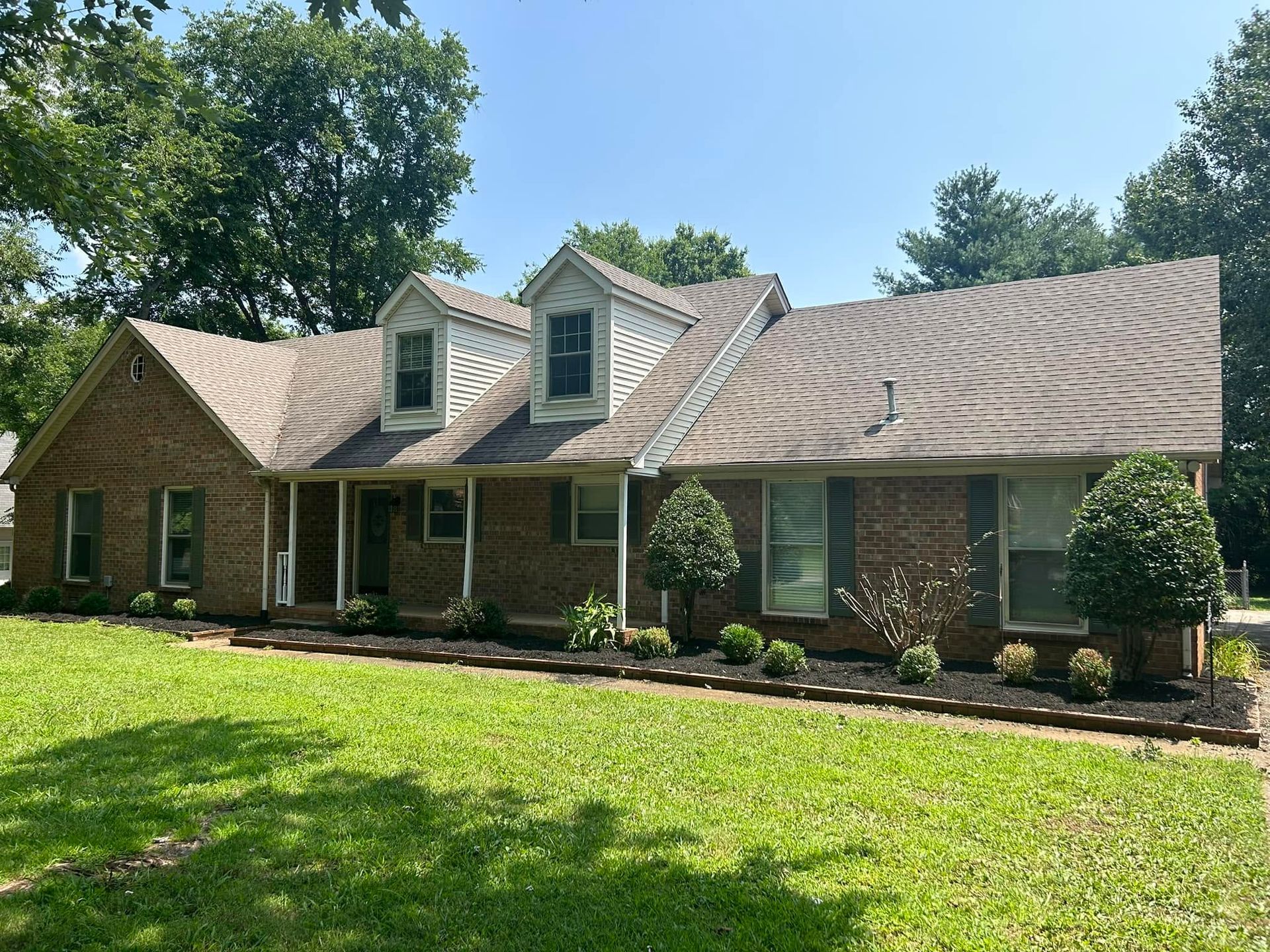Brick suburban house with dormer windows, front porch, and green lawn under a clear blue sky