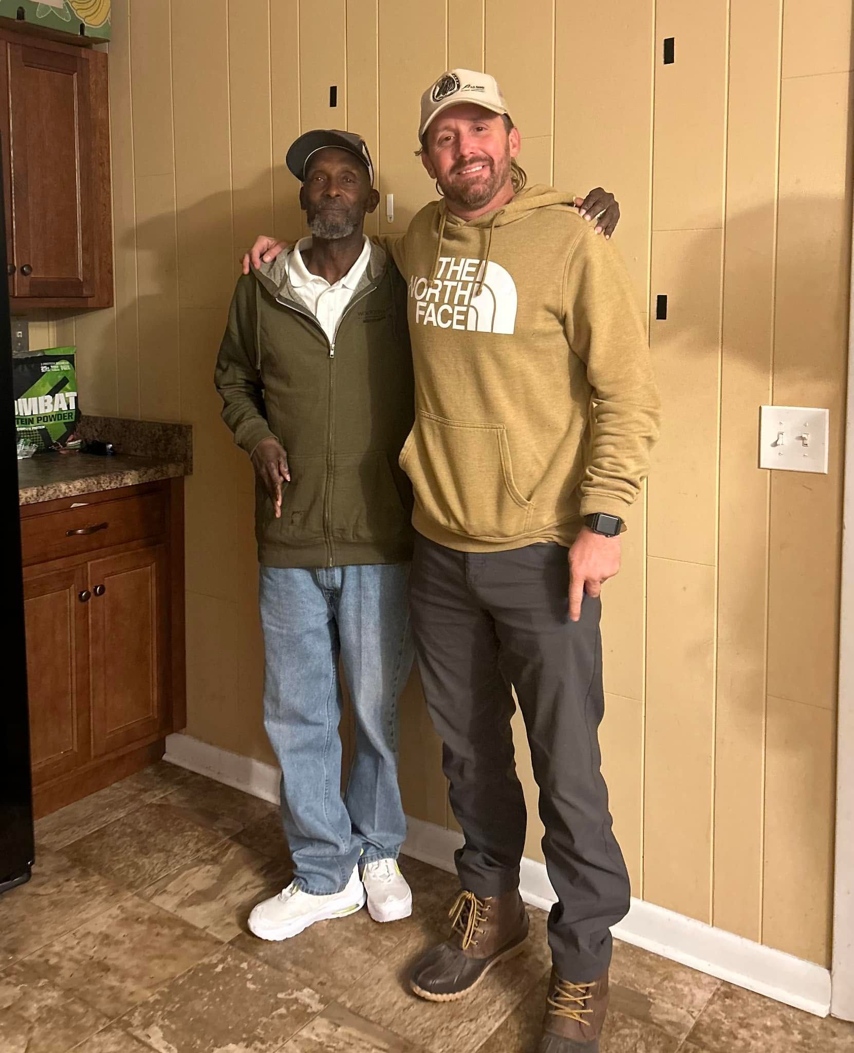 Two people stand together in a kitchen, smiling, wearing casual clothes and boots.