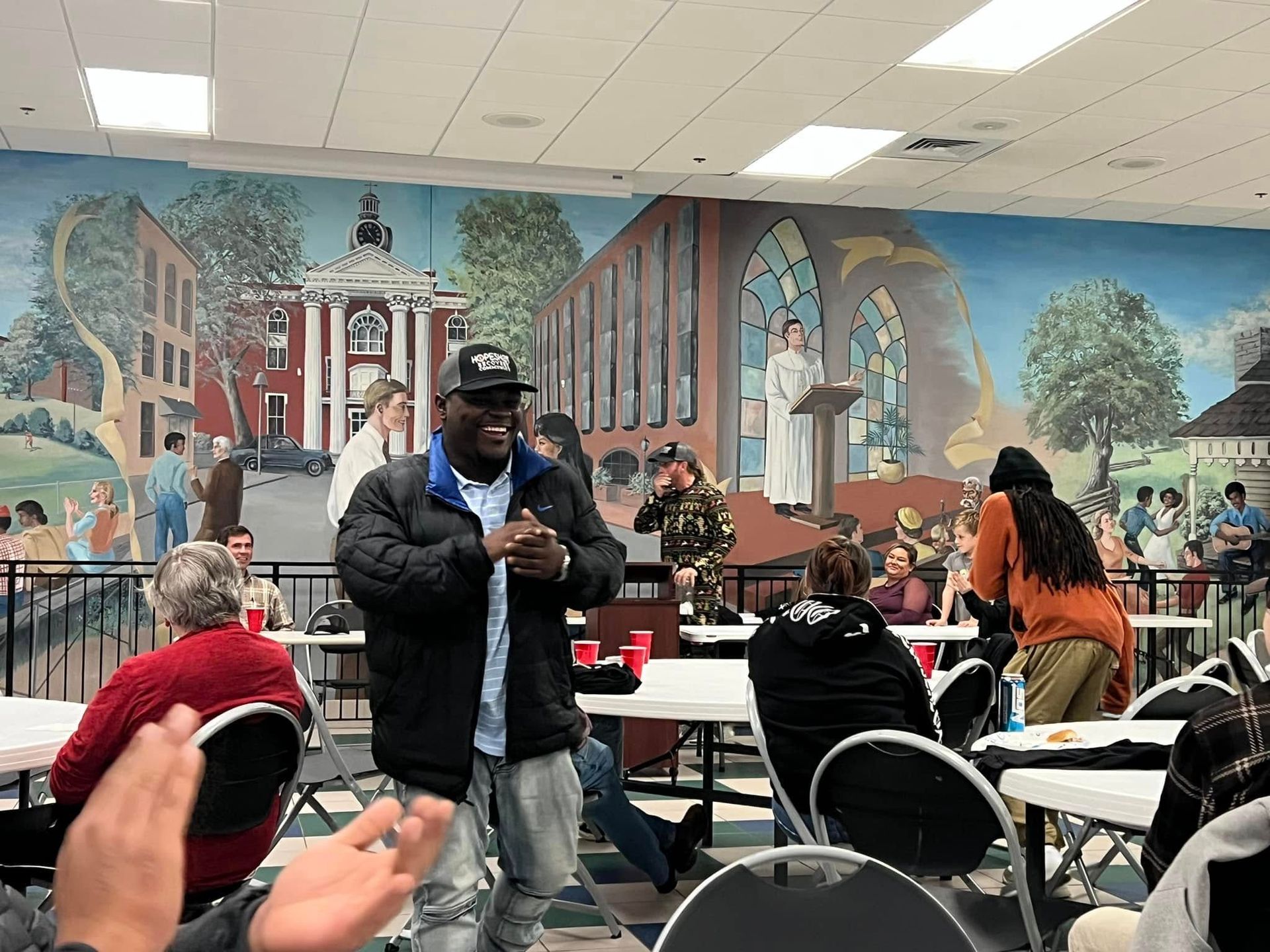 People gathered in a community room with a colorful mural, seated at tables and mingling.