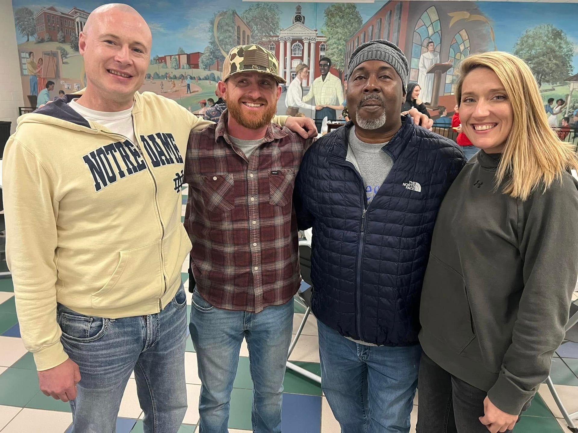 Four people smiling indoors, standing arm in arm in front of a mural.