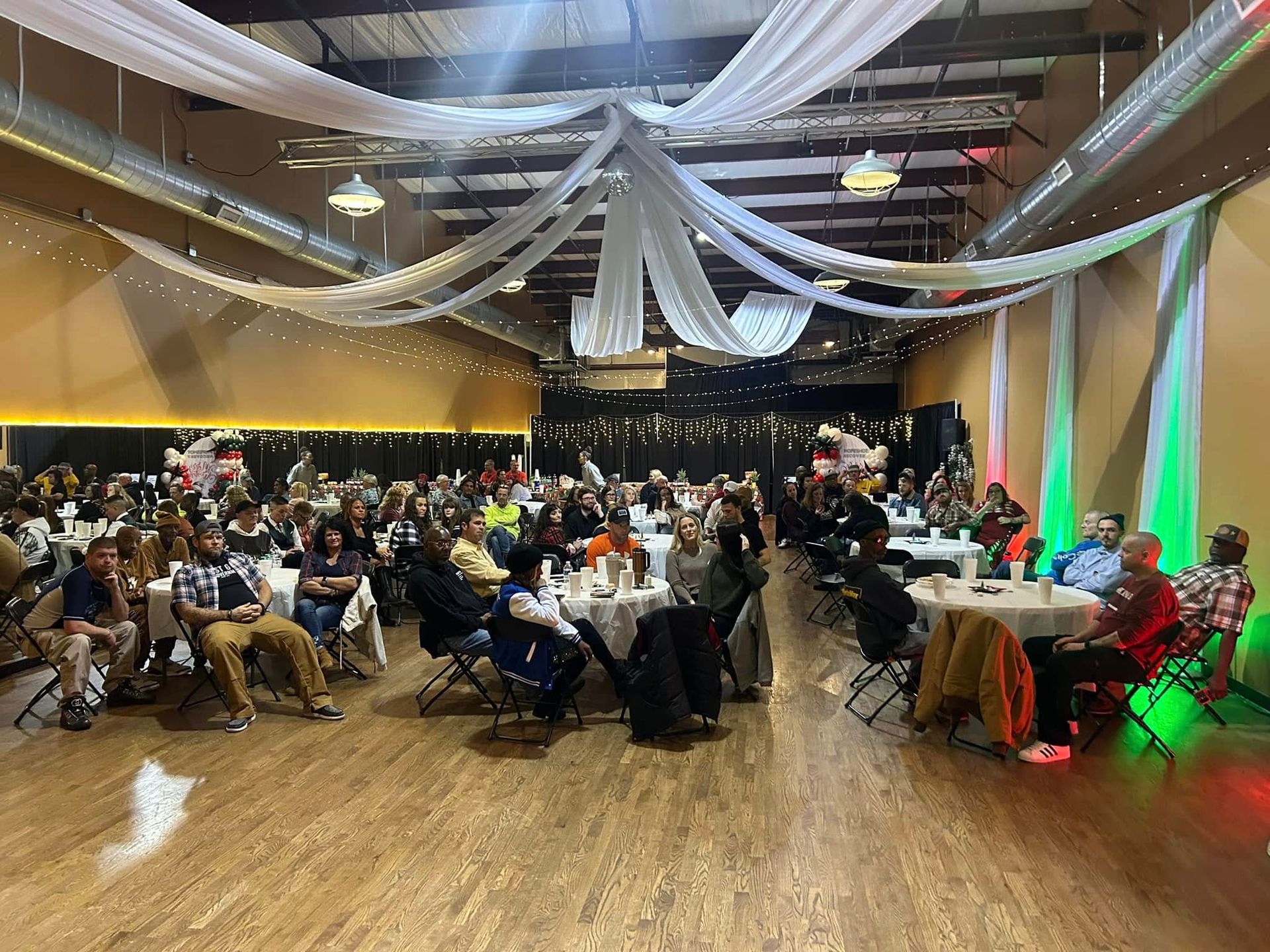 Large banquet hall with many seated guests at round tables under draped ceiling fabric and colored stage lights