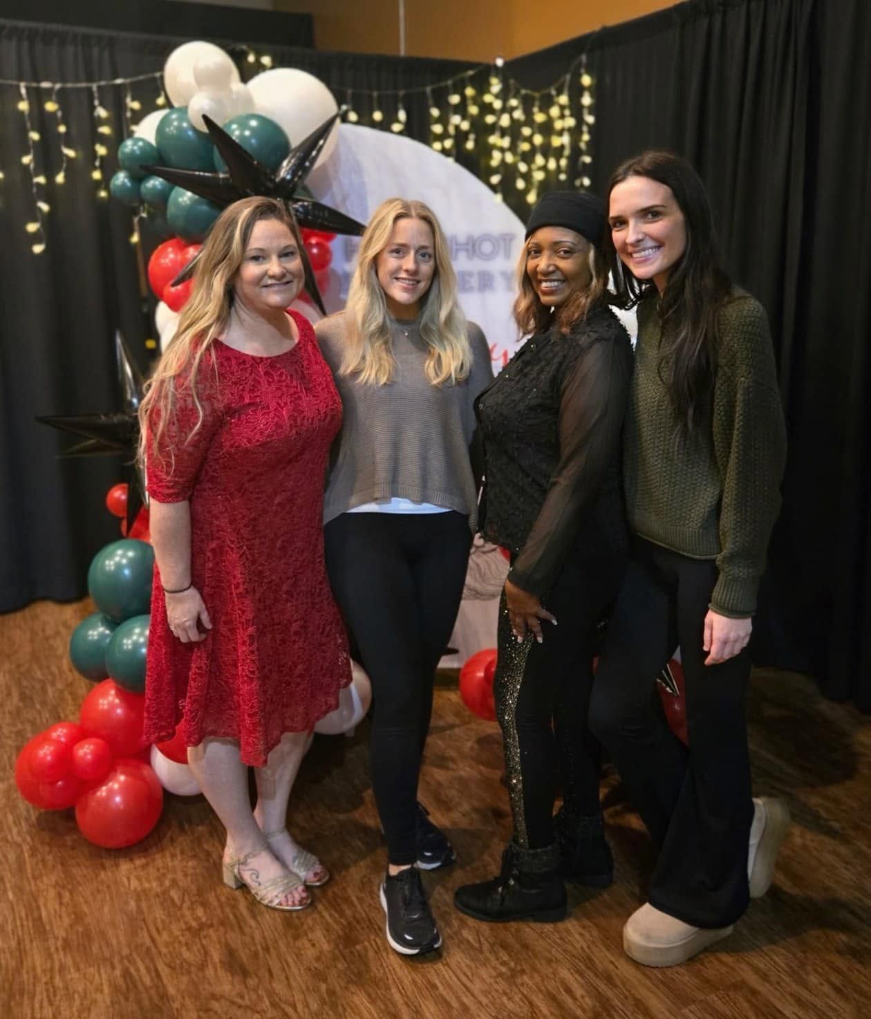 Four women posing at a festive party with balloons and string lights, smiling in front of a decorated backdrop