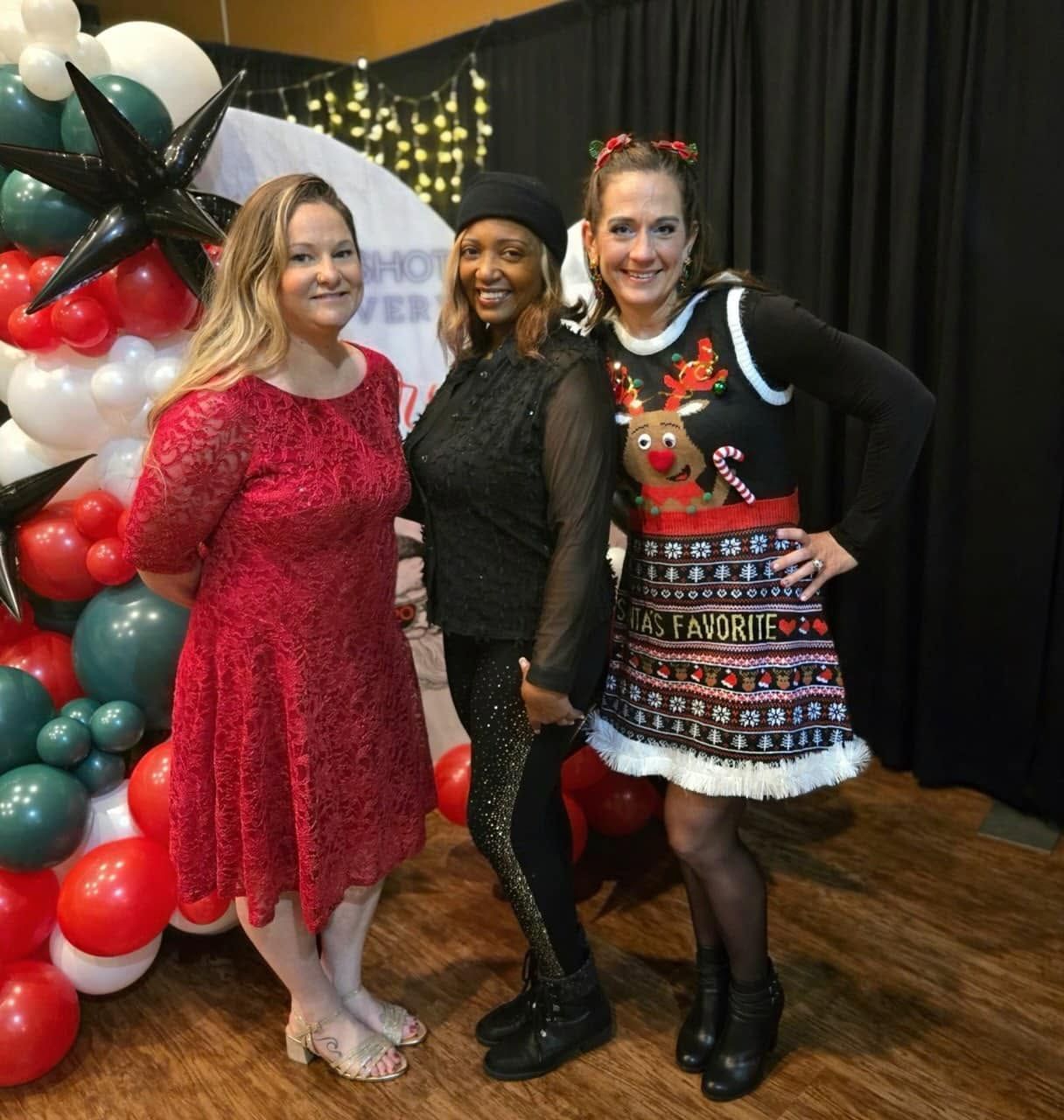 Three women posing at a festive holiday event beside a red, white, and green balloon display