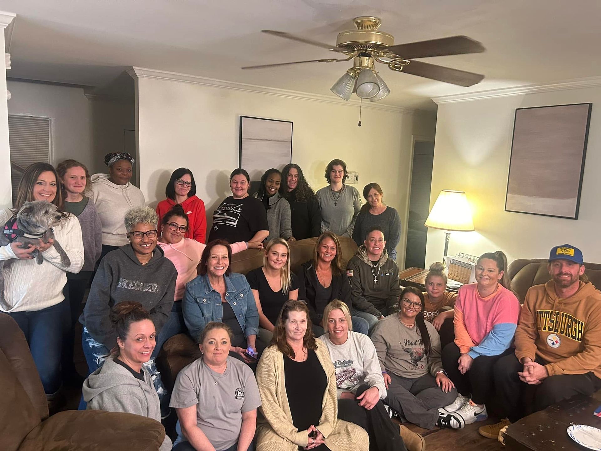 Large group posing together in a living room, seated and standing around a couch under a ceiling fan.