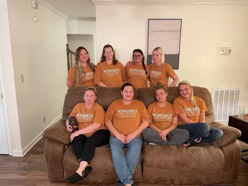 Seven people in orange shirts sit and stand on a brown couch in a living room.