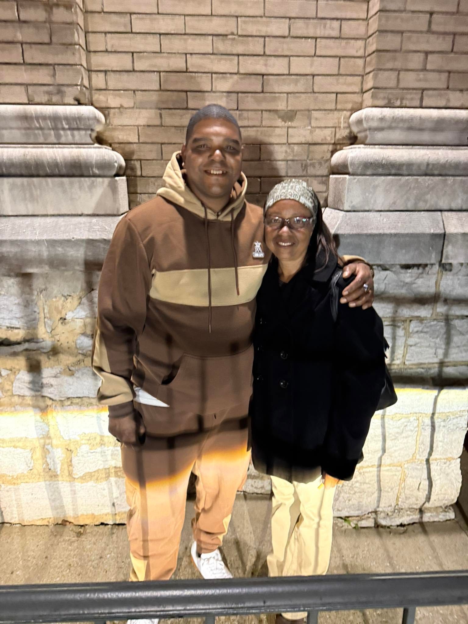 Two people smiling and posing arm-in-arm in front of a stone wall at night.