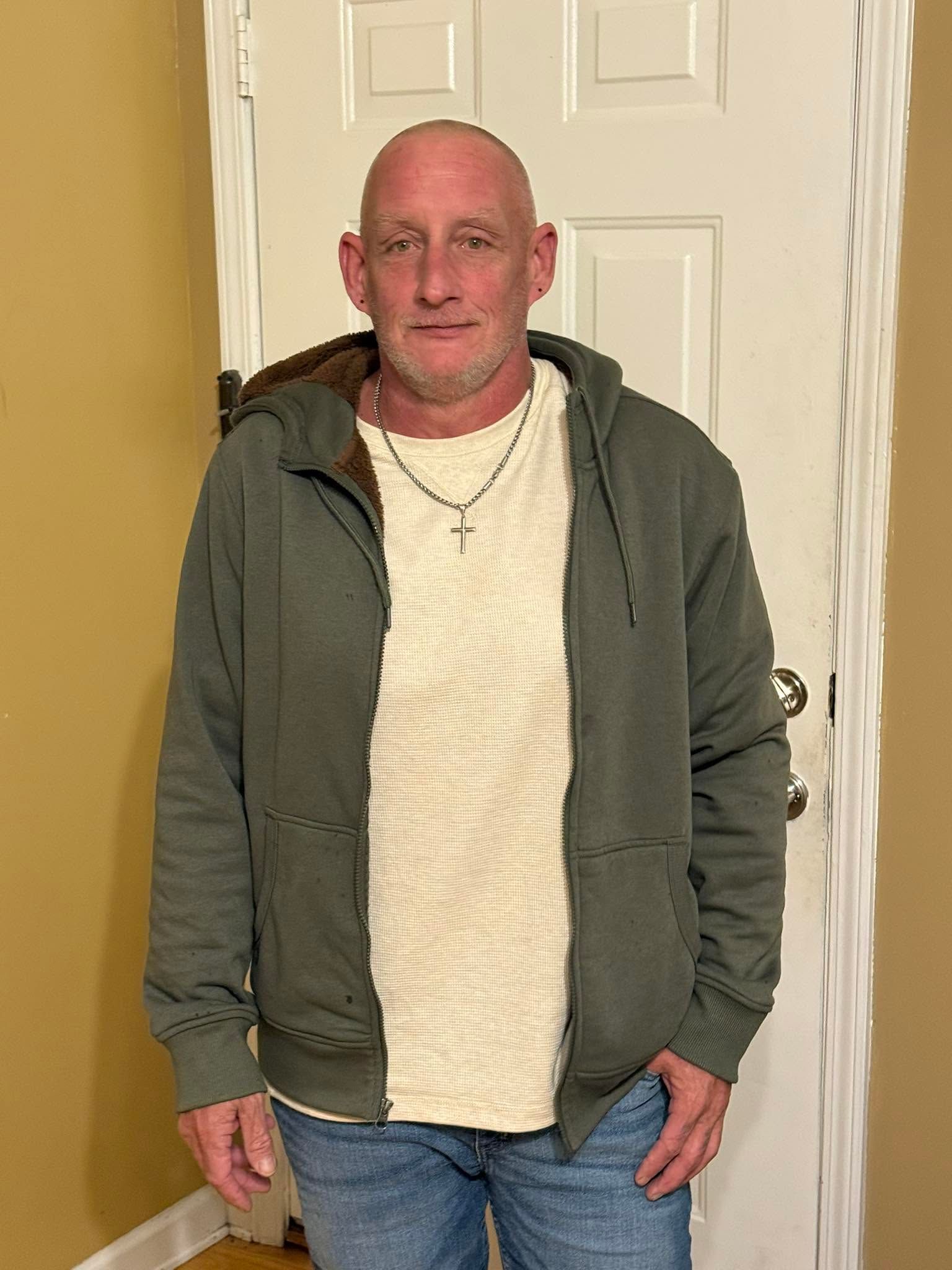 Person standing indoors in a green hoodie and cream sweater in front of a white door.