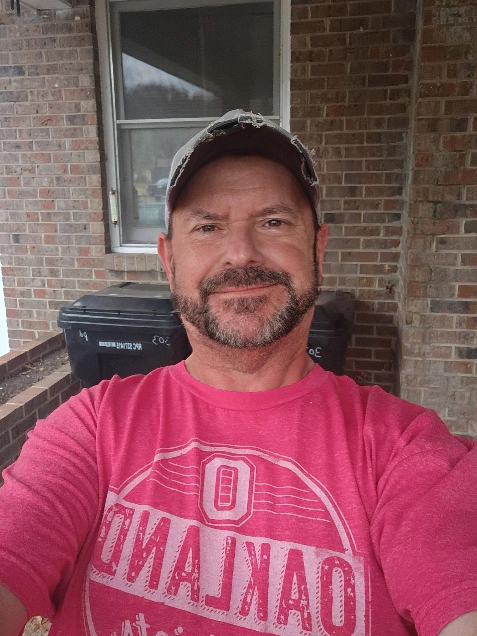 Man in a pink shirt and cap smiling in front of a brick wall and window on a porch