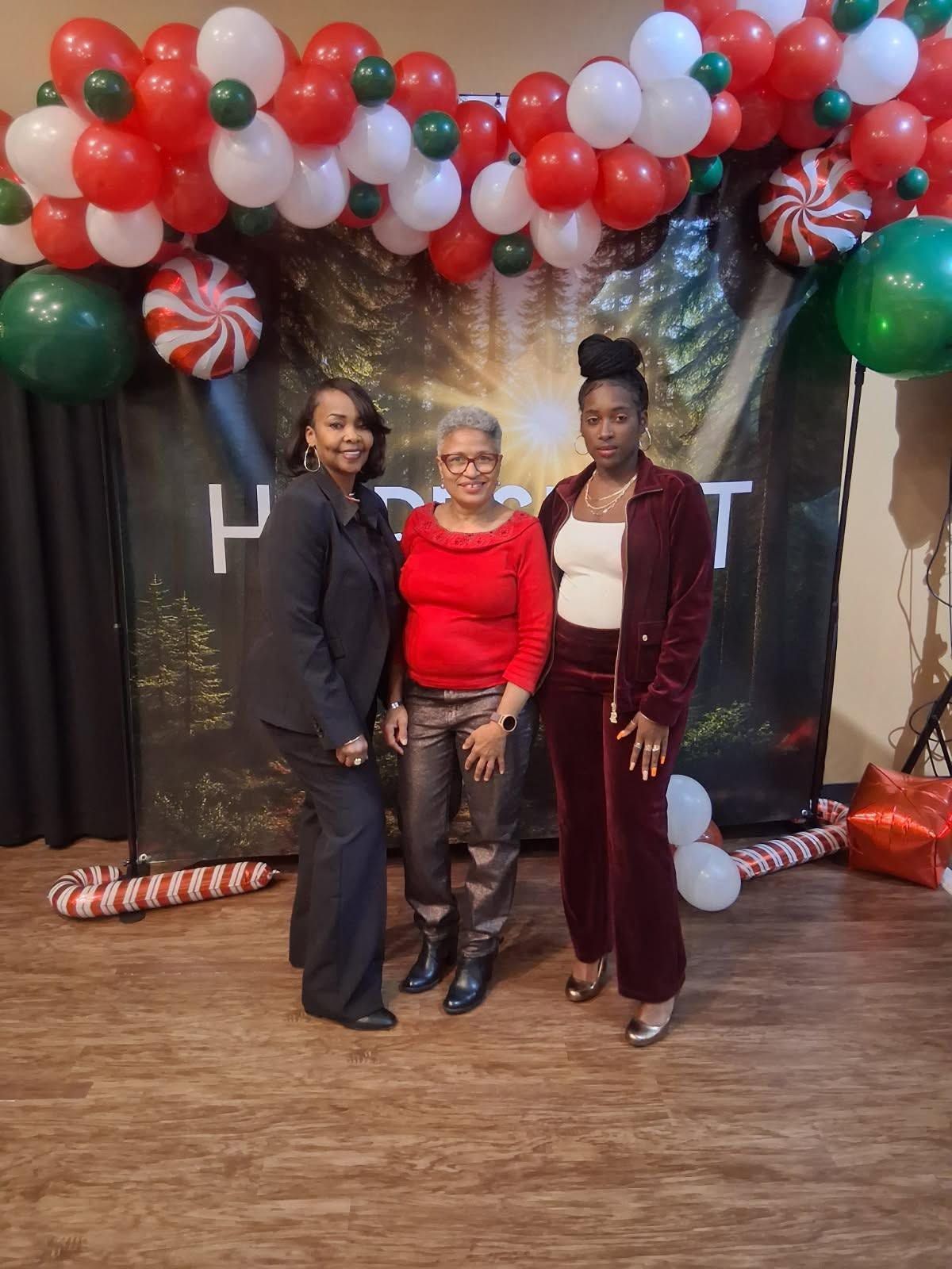 Three people pose under a red, green, and white balloon arch in front of a black backdrop.