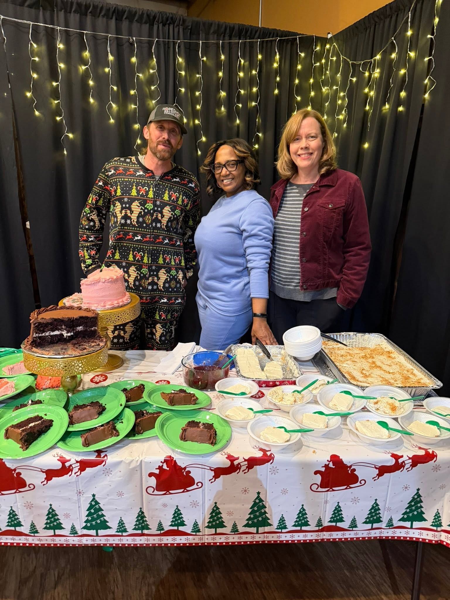 Three people behind a dessert table with green-frosted cookies and a cake, in front of black curtains with string lights