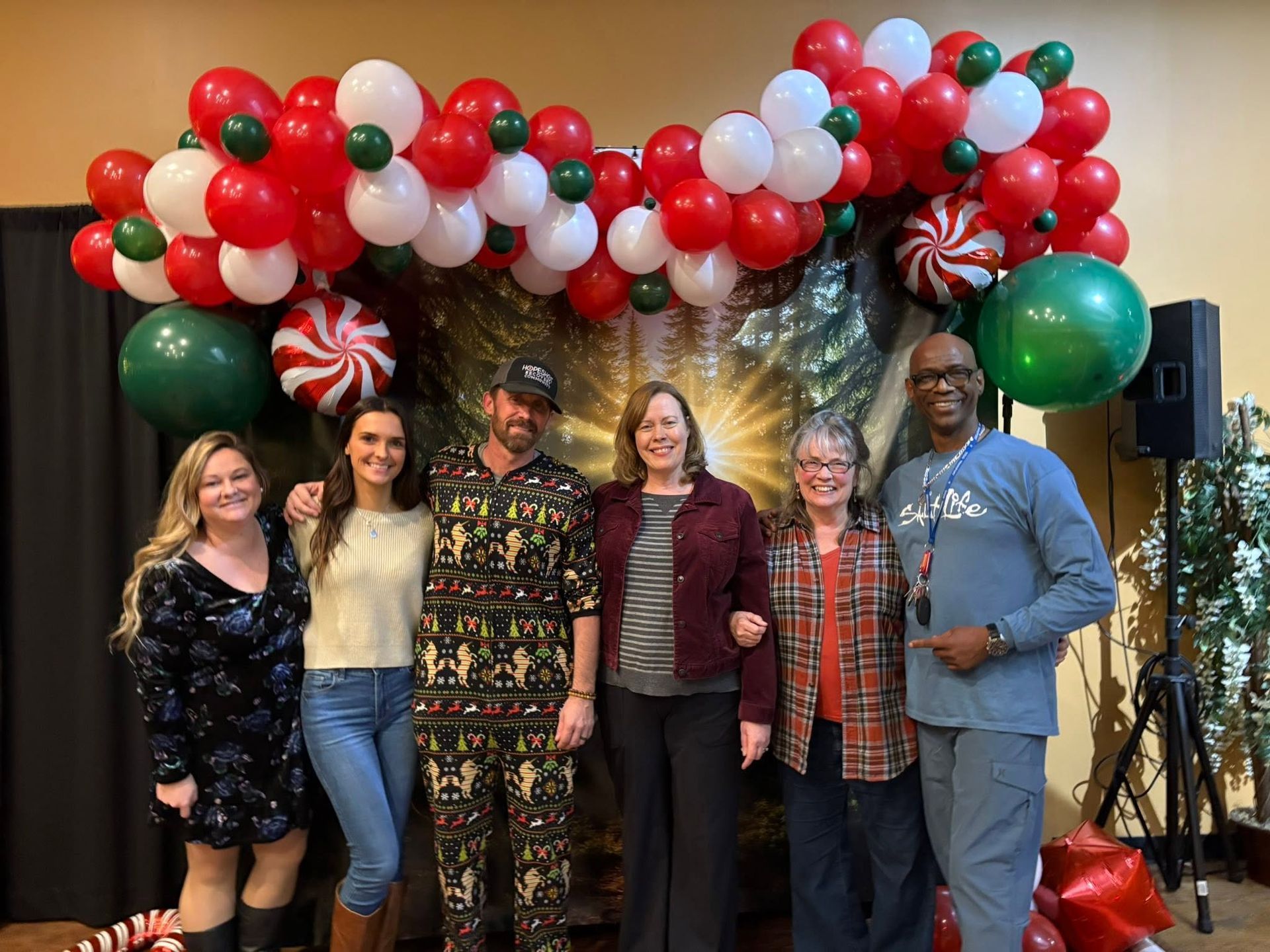 Six people posing under a red, white, and green balloon arch at an indoor event backdrop.