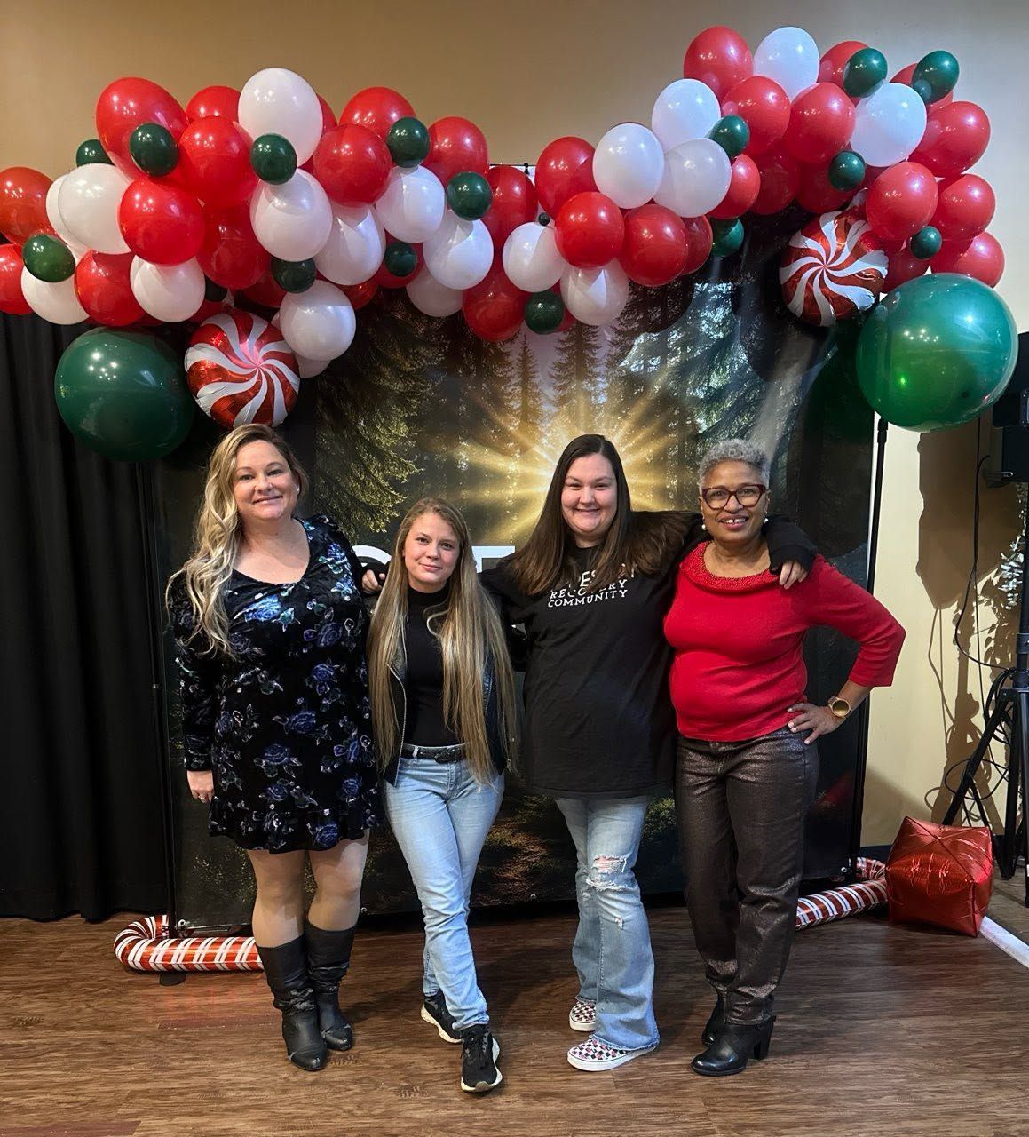 Four people pose indoors under a red, white, and green balloon arch.