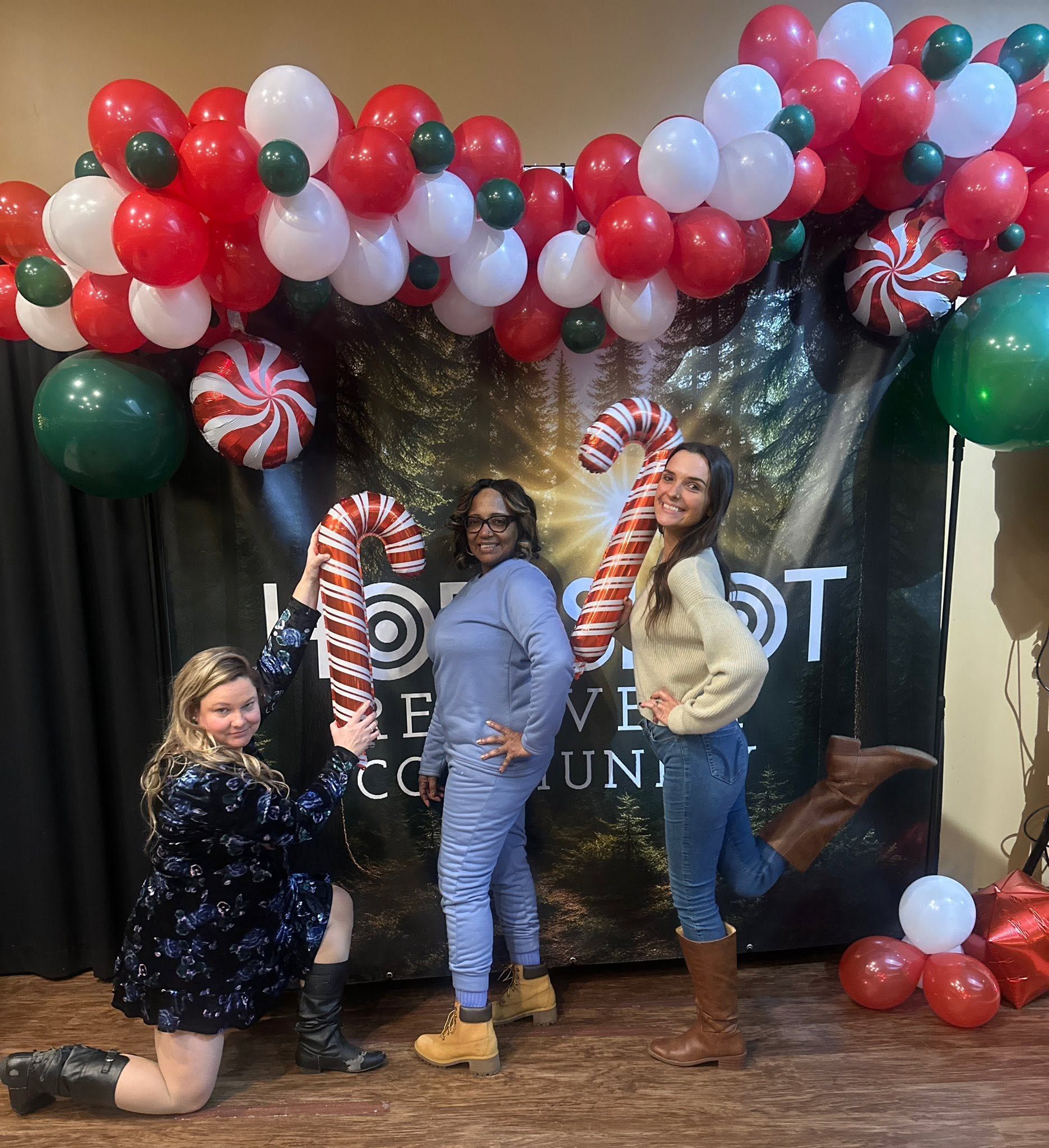Three women pose with candy canes under red, white, and green balloon arch at an event backdrop.