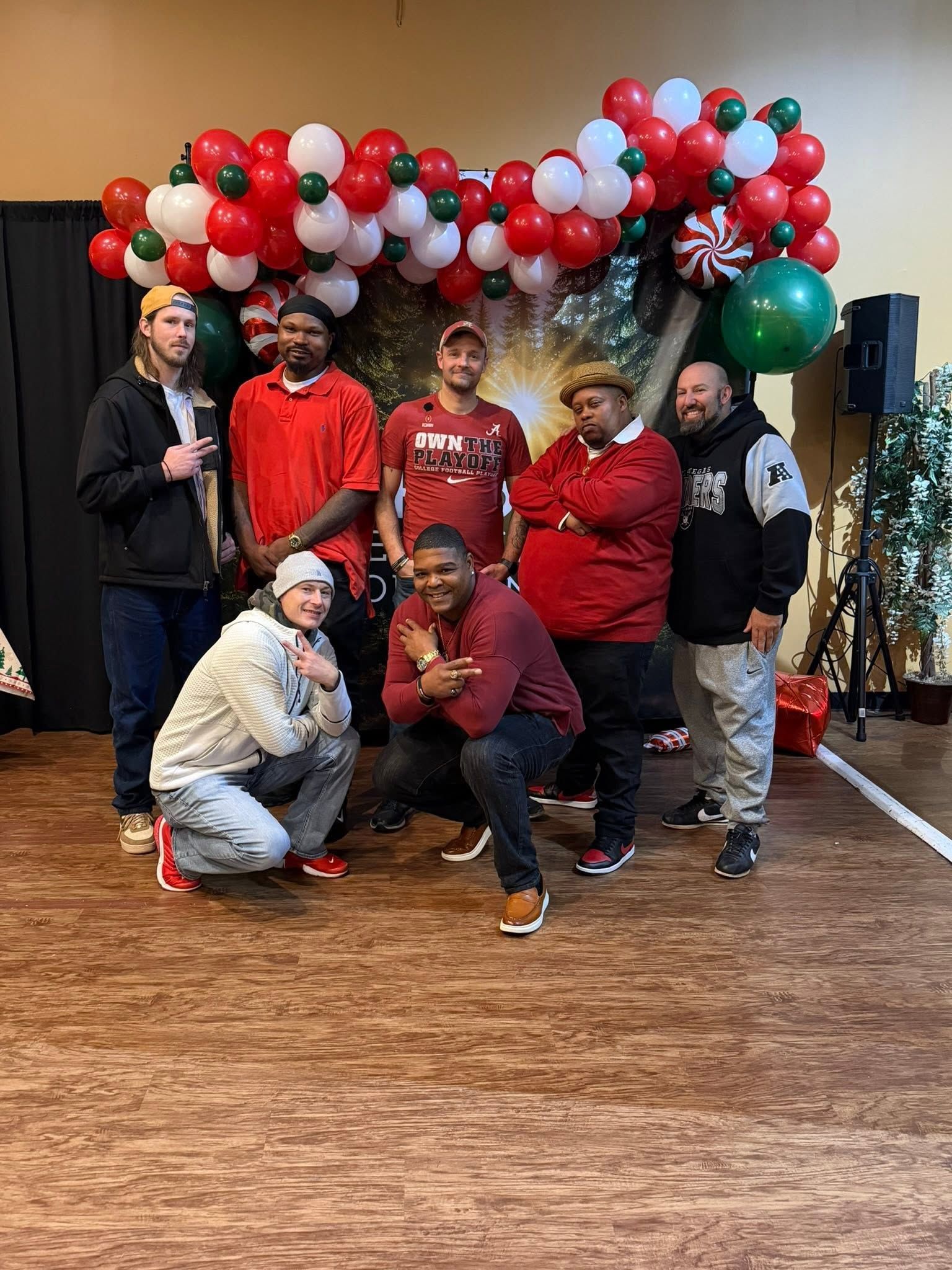 Group posing under a red, white, and green balloon arch at an indoor celebration.