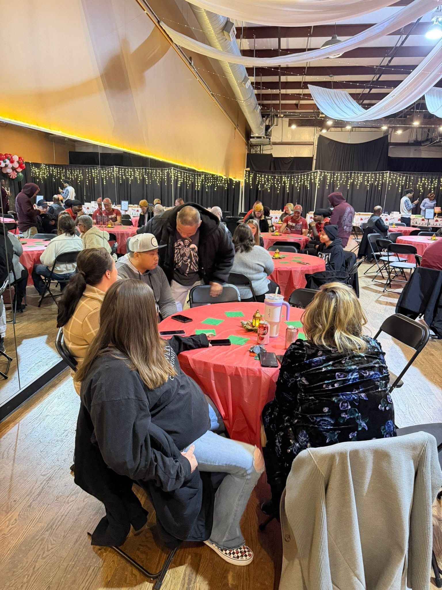 Indoor banquet hall with people seated at red-covered tables under draped ceiling lights