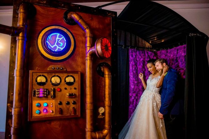 Steampunk-style photo booth beside a bride and groom posing under purple lights and a glittery backdrop