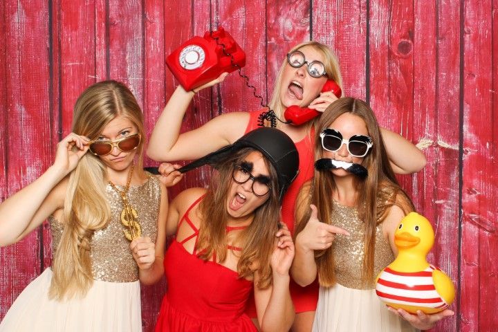 Five women posing with props in front of a red wooden backdrop, wearing sunglasses and playful expressions