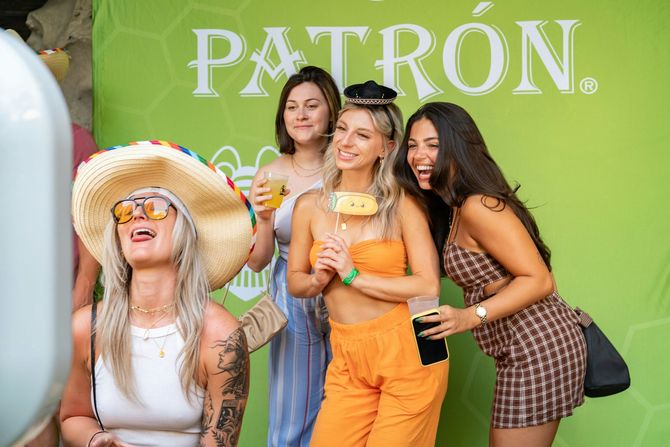 Three women posing at a Patrón-branded event backdrop, smiling and holding drinks