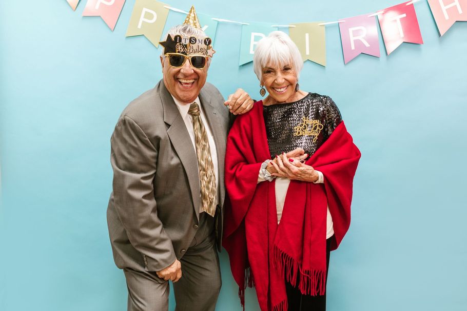 Two smiling people posing in front of a birthday banner, one wearing a gold crown and the other a red shawl