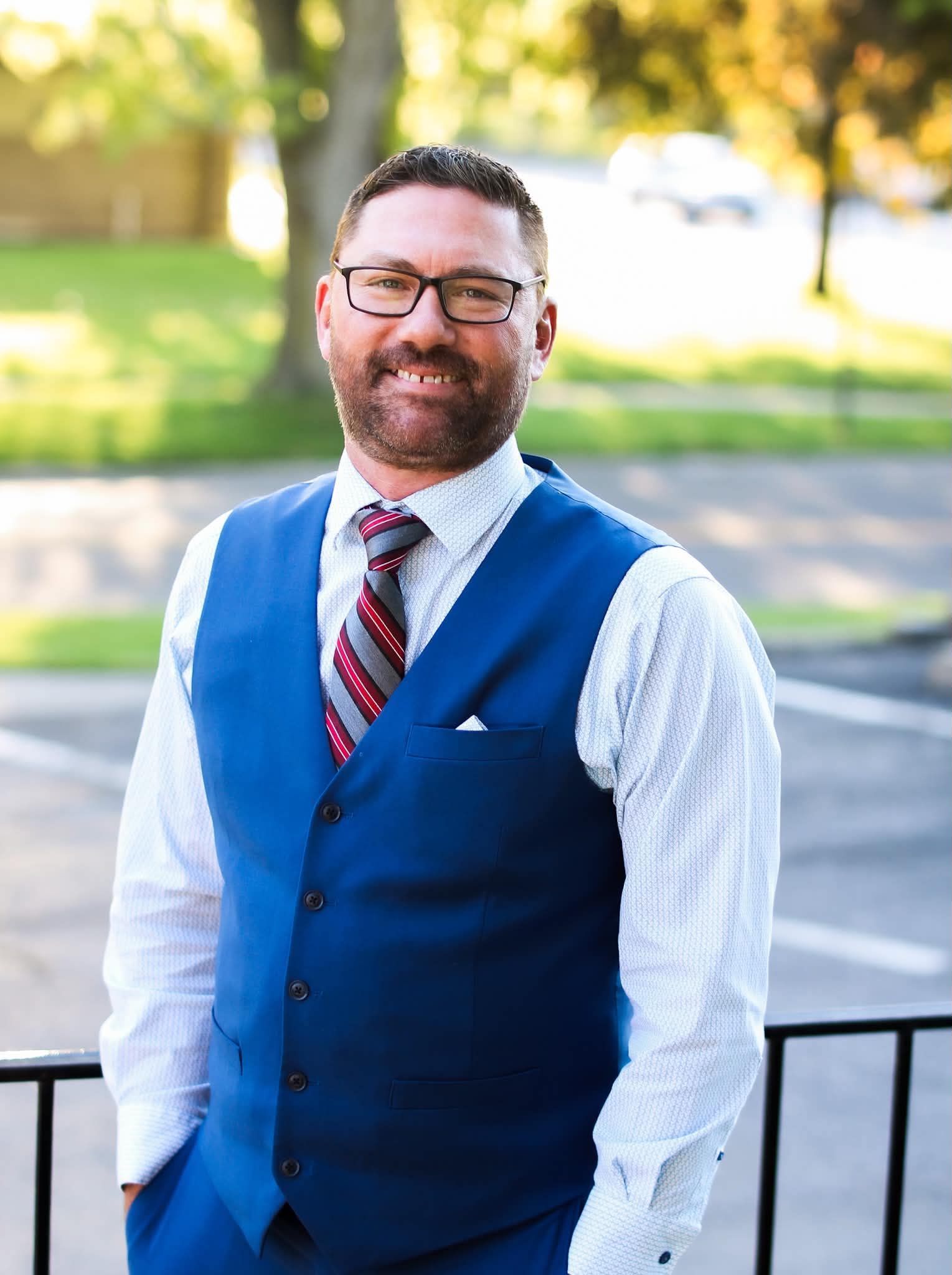 Man in blue vest and striped tie standing outdoors by a railing, smiling in a sunlit park.