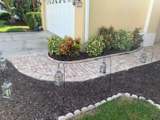 A curved brick walkway beside a house, lined with decorative hanging lanterns and low-maintenance landscaping.