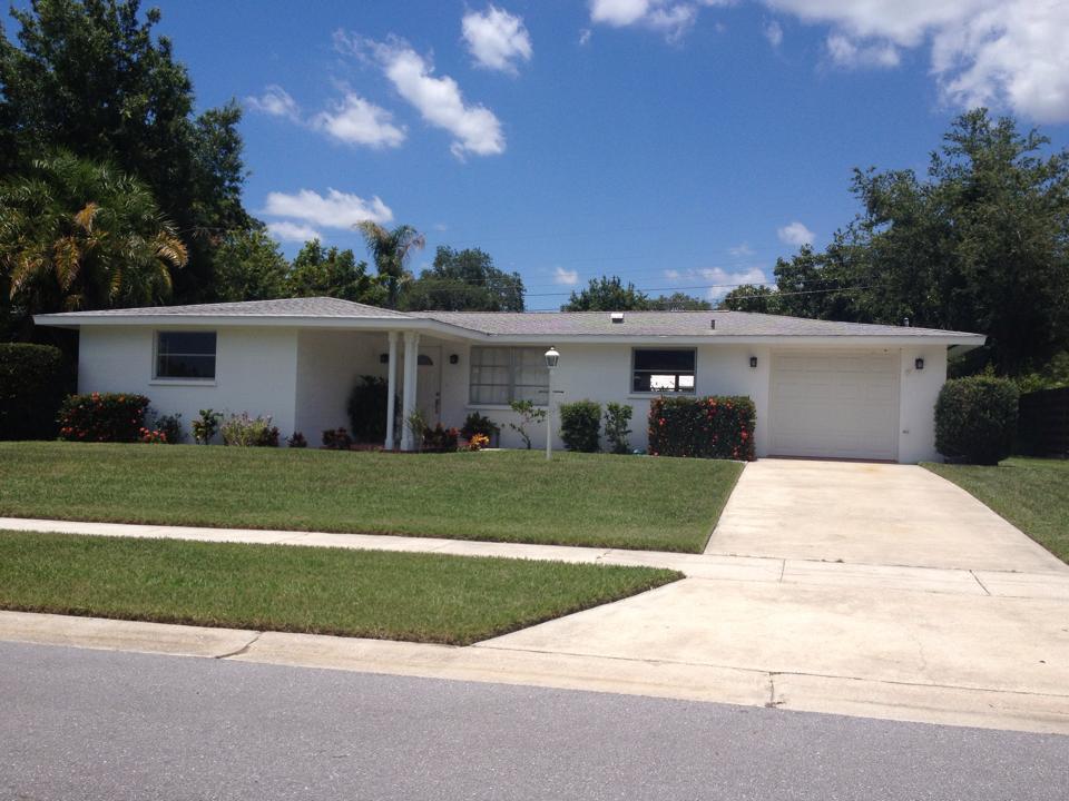 A white, single-story ranch-style house with a driveway and a neatly manicured lawn under a sunny, blue sky.